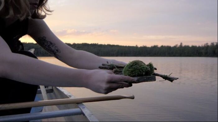 A screenshot from the short film Wolf Inventory featuring a trans femme lowering a mossy assemblage over the side of a rowboat.